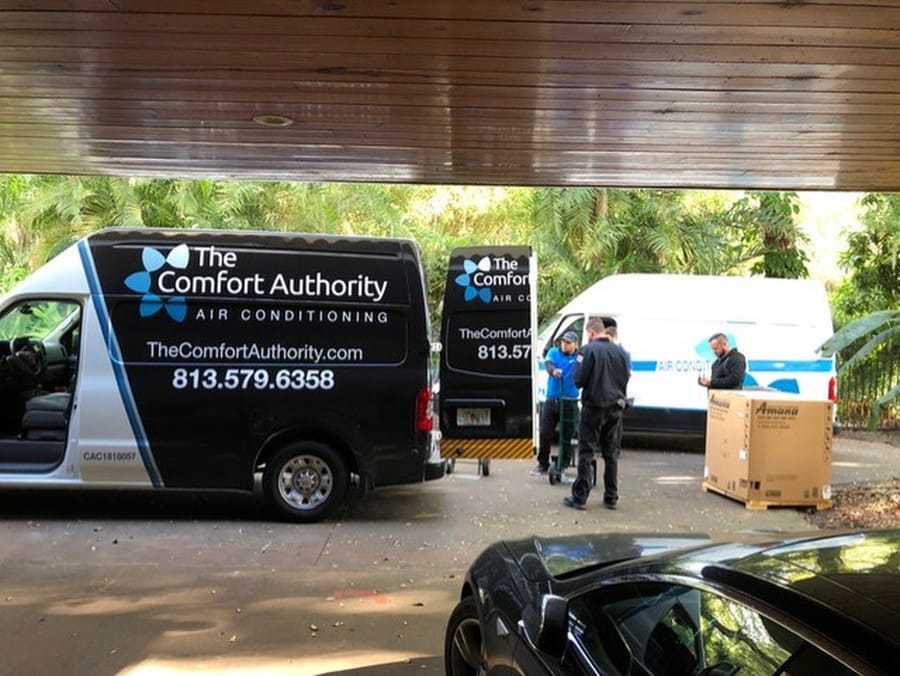 Two service vans from The Comfort Authority, are parked in a driveway surrounded by lush greenery. Three technicians are visible, engaged in a discussion about the repair service.