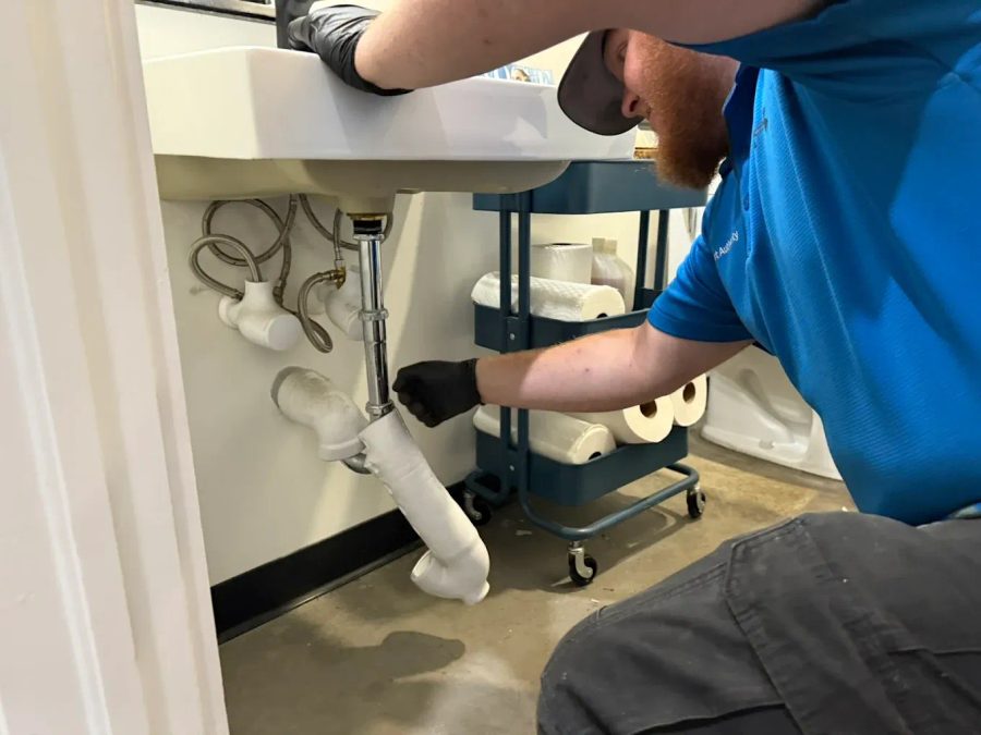 Plumber kneeling under a sink, adjusting white PVC pipes with a wrench, wearing a blue shirt and gloves.